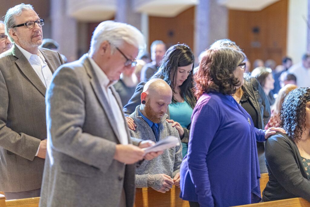 Man kneels, with godfather's hand on shoulder during the Rite of Election. Photo by Brian Keith.