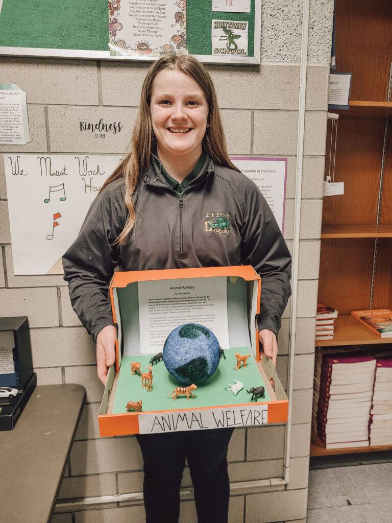 Student smiles, holding diorama featuring paper mache planet.