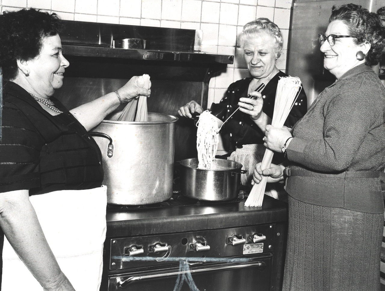 Black and white image of three women standing around a large pot of spaghetti