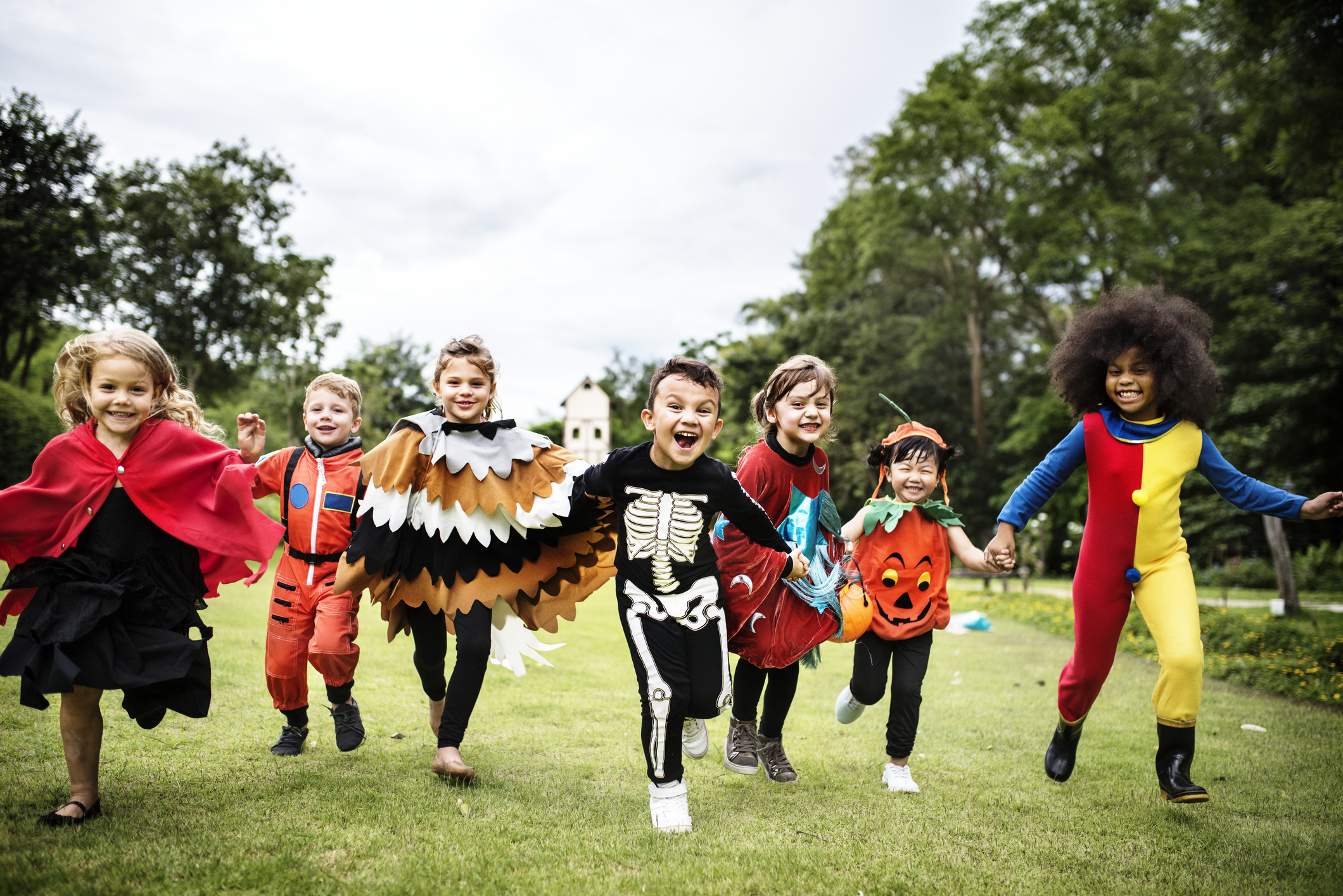 A group of kids runnning through a park dressed up in Halloween costumes.