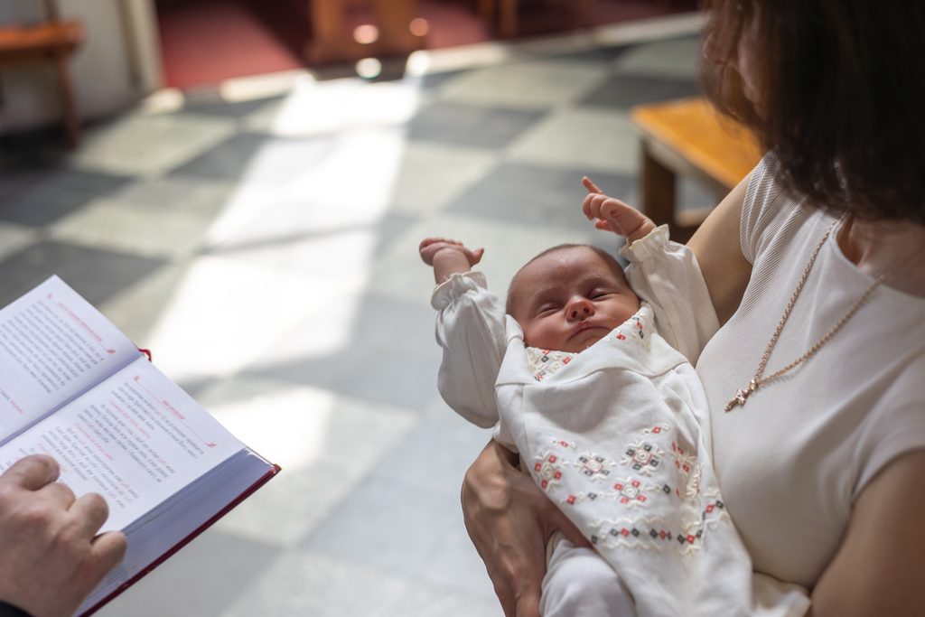 A newborn baby in a white embroidered christening robe sleeps in a woman's arms during a baptism ceremony in a sunlit church, while a priest holds open a prayer book — the kind of quiet, sacred moment church cry rooms are designed to protect.