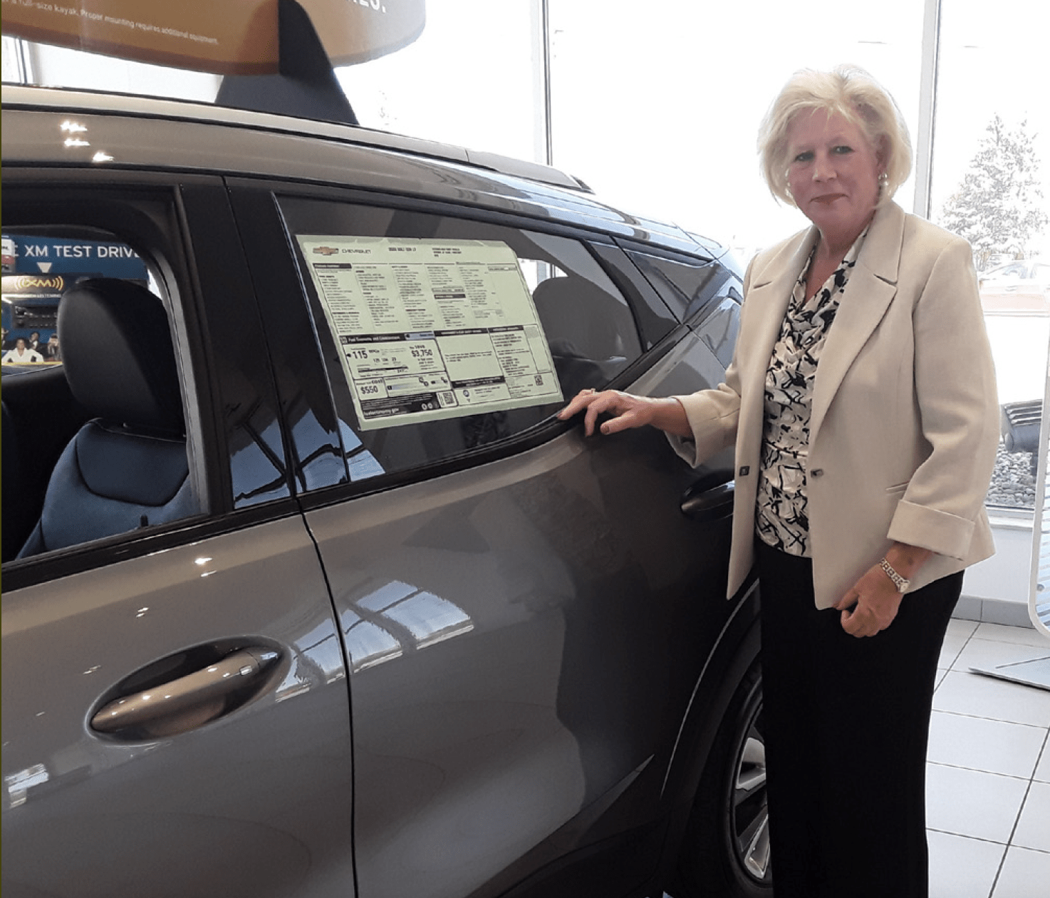 Diane Sauer stands in front of a silver car in her car dealership.