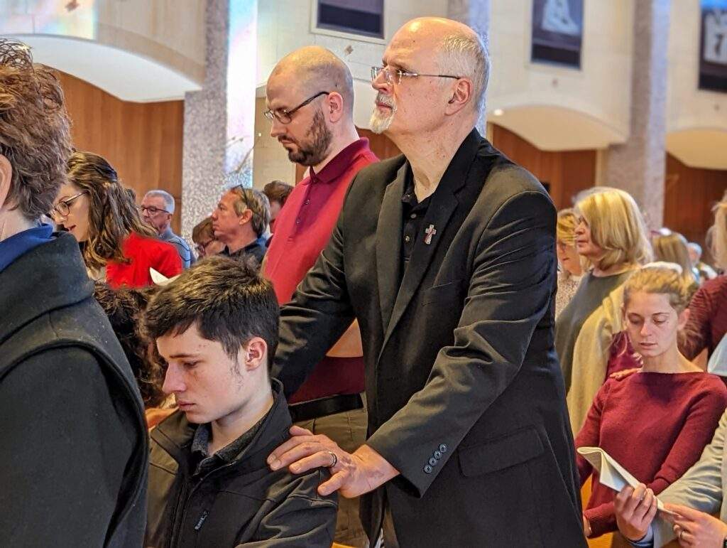 A godparent stands with his hands on the shoulders of a young catechumen.