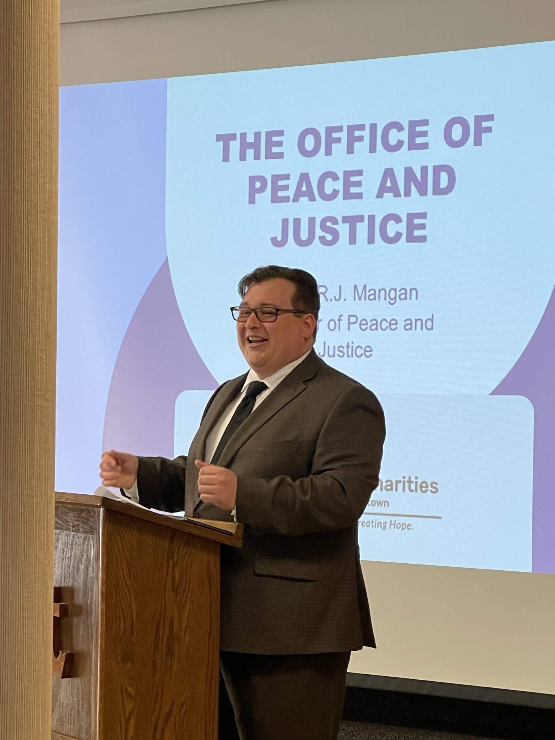 R.J. Mangan, Catholic Charities' Director of Peace and Justice, stands at a podium and runs a presentation.