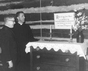 Two priests look at a sign that reads "Altar of our First Mass 1817 celebrated by the Rev. Edward Fenwick D.D. First Bishop of Cincinnati and First Pastor of Dungannon" hung on top of a dresser that was once used as an altar.