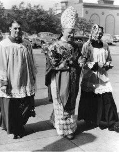 Bishop McFadden, in full vestments and holding a crozier, crosses a road in Youngstown, flanked by two priests.