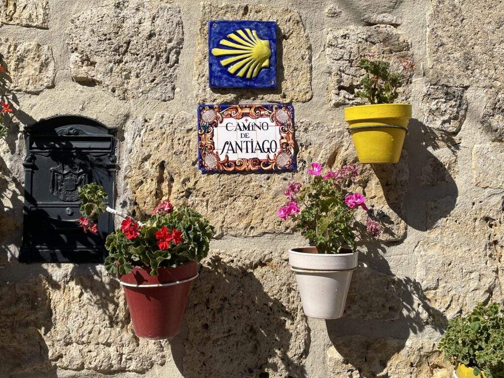 Two sets of tiles featuring the Camino scallop shell and the words "Camino de Santiago," surrounded by colorful flower pots.