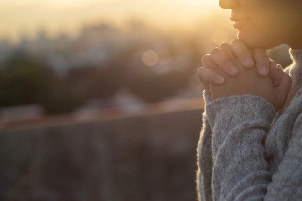 Woman holds hands up in prayer, while the sun sets behind her.