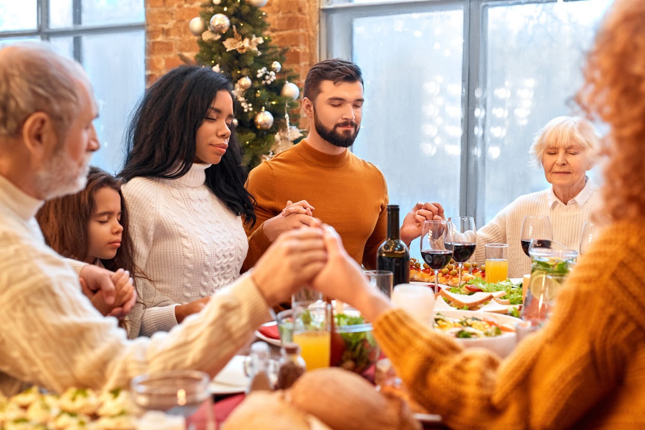 Mixed race family holding hands and praying with eyes closed before vegan holiday dinner at home. Christmas, New year, Thanksgiving, Anniversary, Hanukkah, Easter, Mothers day celebration concept
