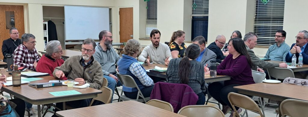 Representatives from 15 parishes and collaborations sit in groups at tables and listen to Ross Bradley from Basilica of St. John and St. Peter Parish in Canton