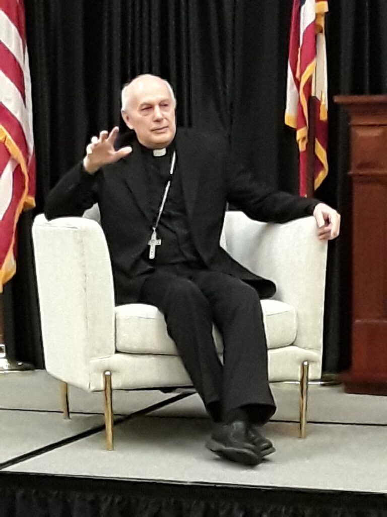 Archbishop, seated in an armchair, speaks to the crowd.