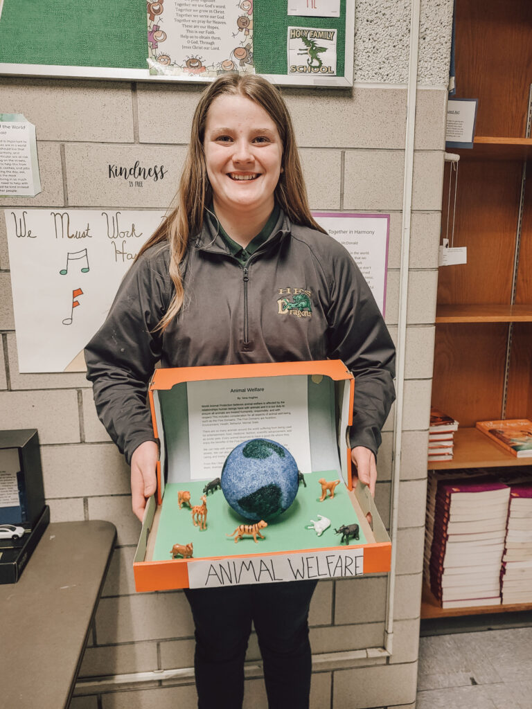 Student smiles, holding diorama featuring paper mache planet.