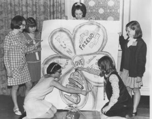 Image of six young women, posing around a large poster of a daisy, which they created.