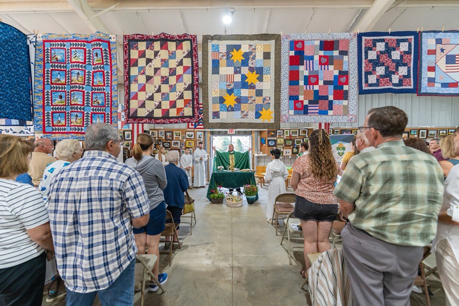 Image of bishop bonnar, praying a Mass inside a fair pavilion, with quilts hanging on the walls behind him