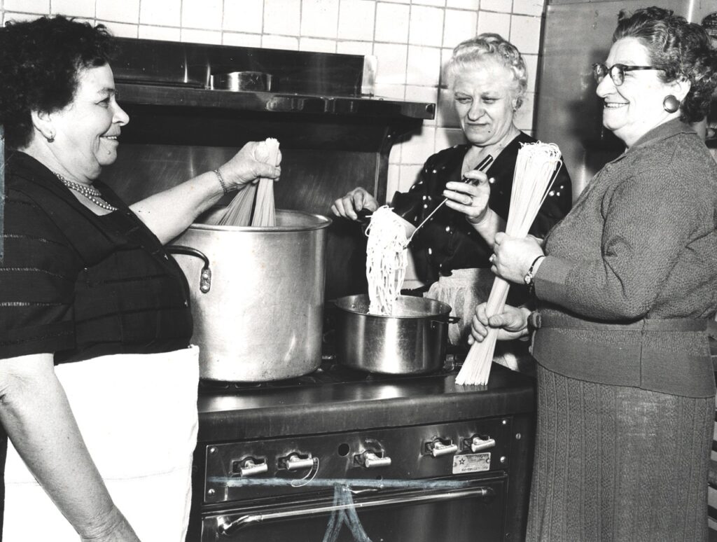 Black and white image of three women standing around a large pot of spaghetti
