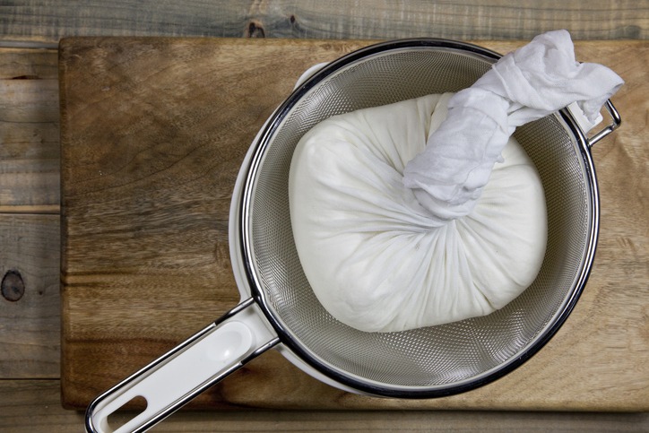 a chunk of cheese squeezed into white bowl through a colander on a rustic wood cutting board after taking the drained whey out.