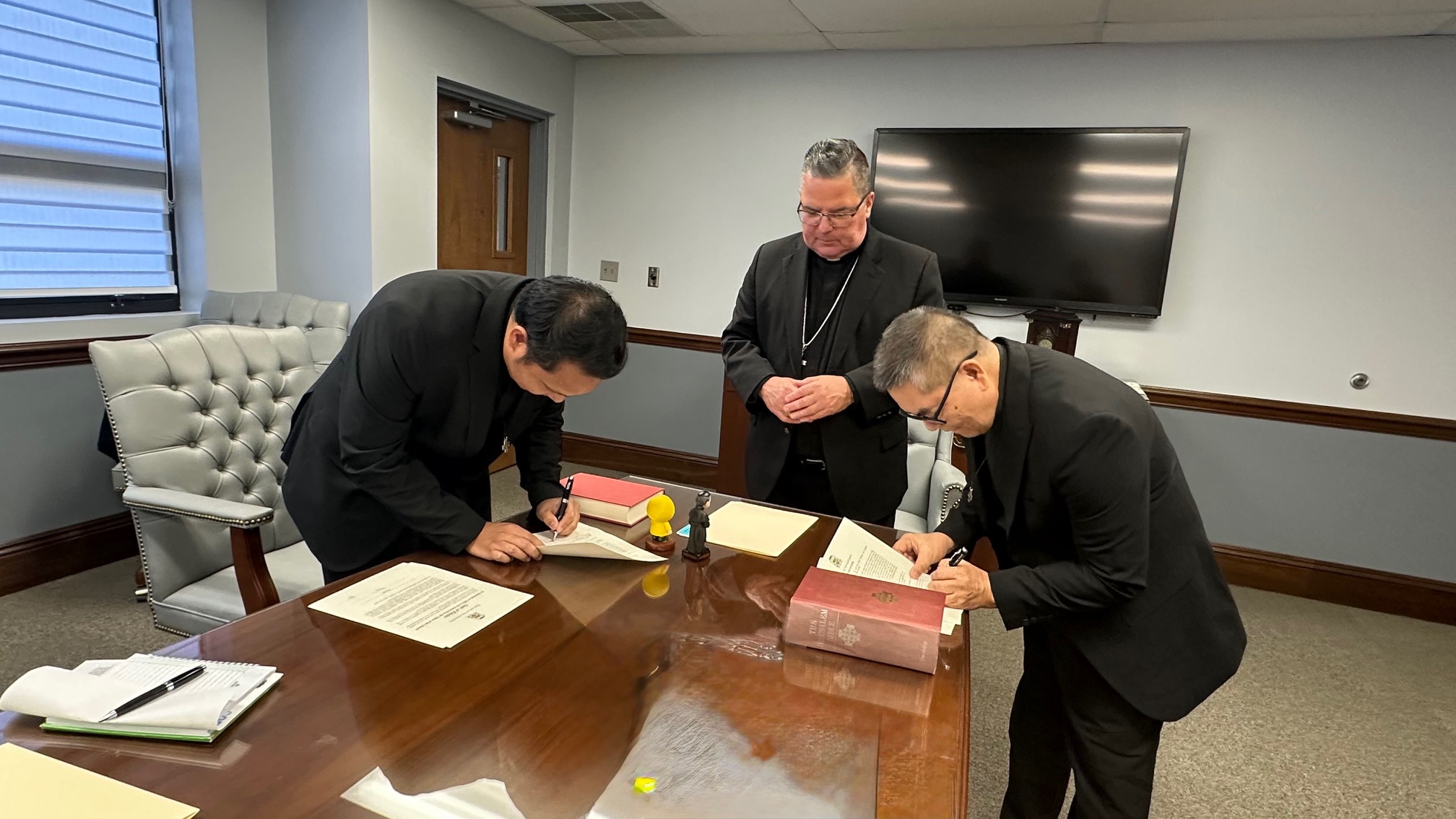Two Rogationists sign a MOU while bishop bonnar watches.